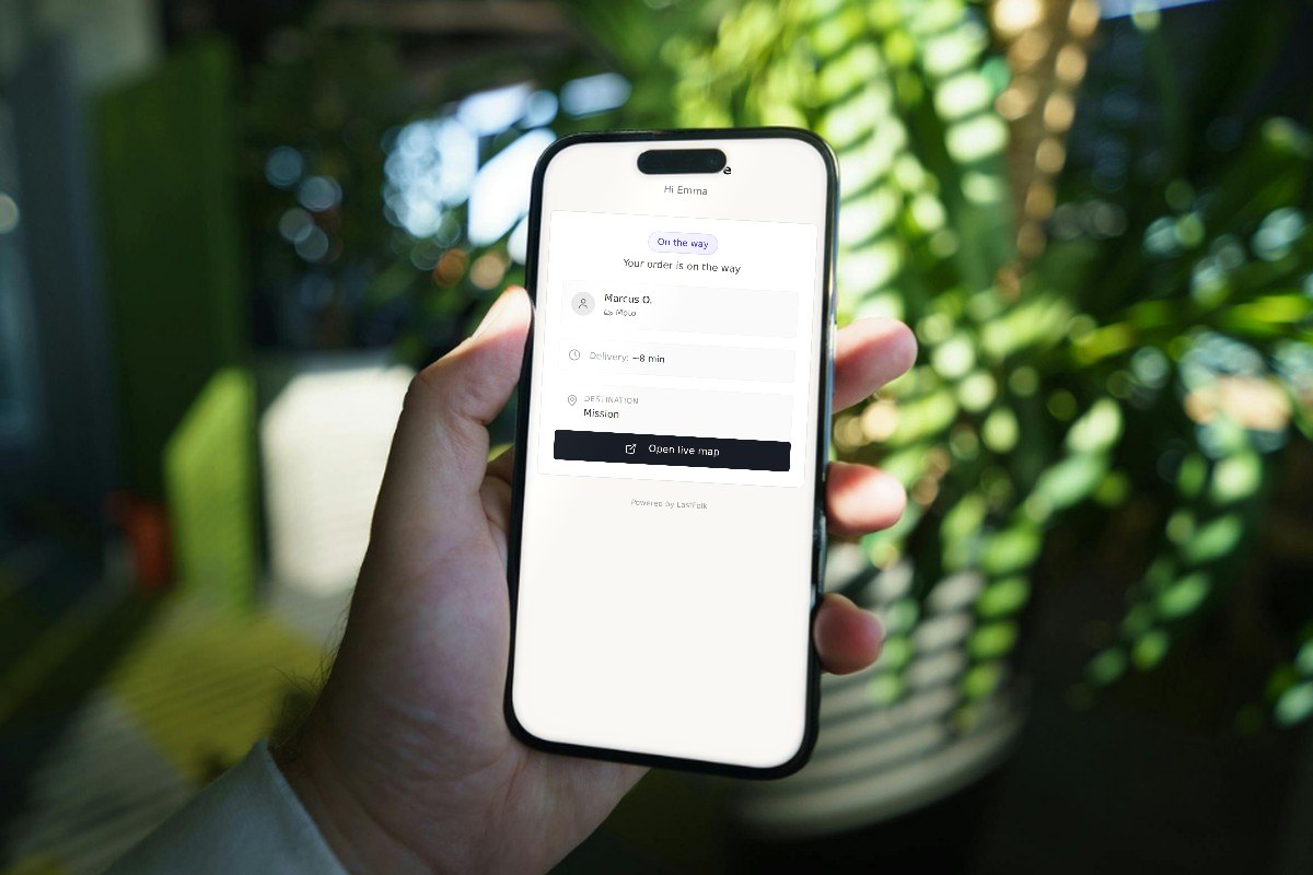 A hand holds a phone showing the tracking page of an order on its way, with demo data, in front of blurred plants.