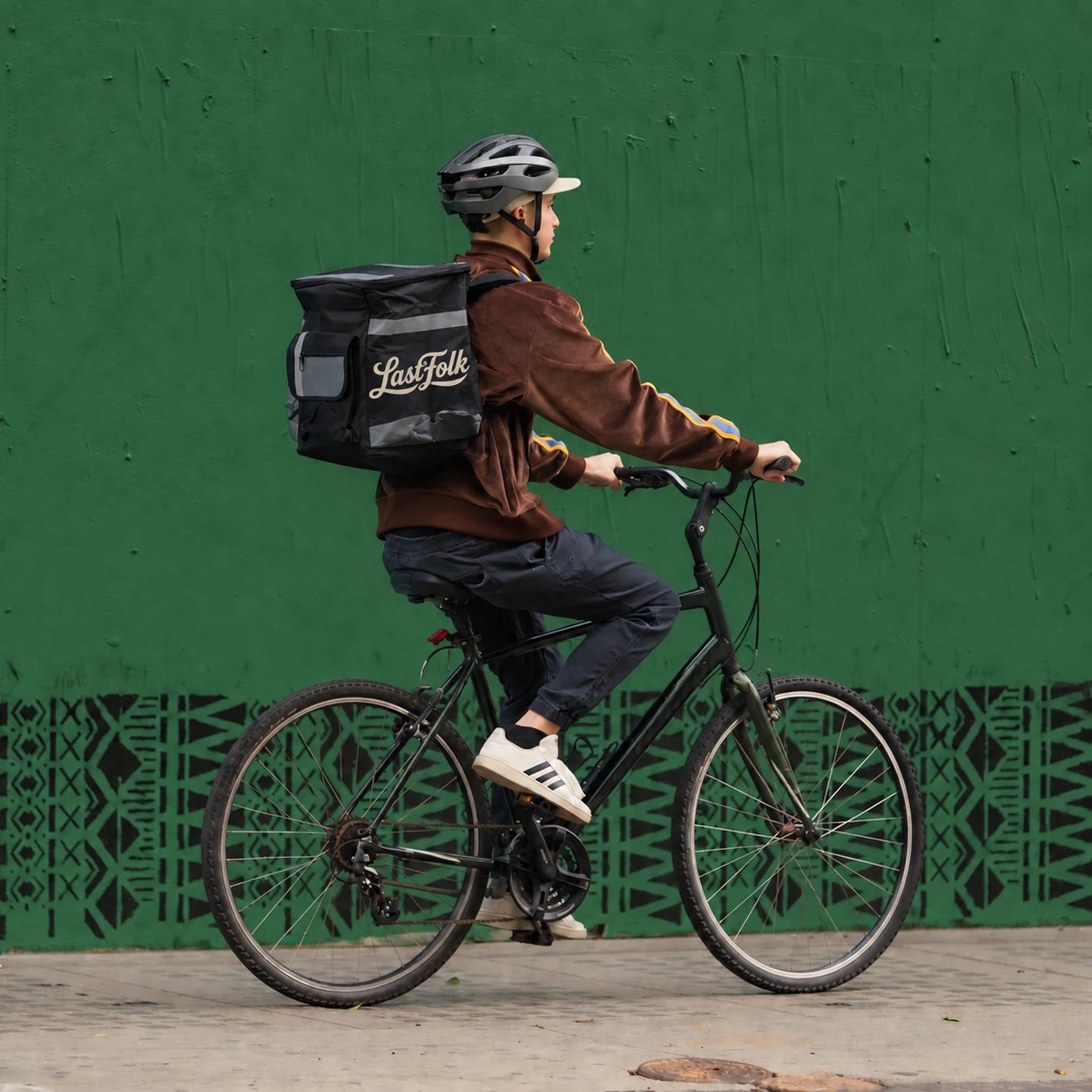 Un repartidor en bicicleta, de perfil frente a un muro verde, con una mochila térmica que lleva el nombre de LastFolk.